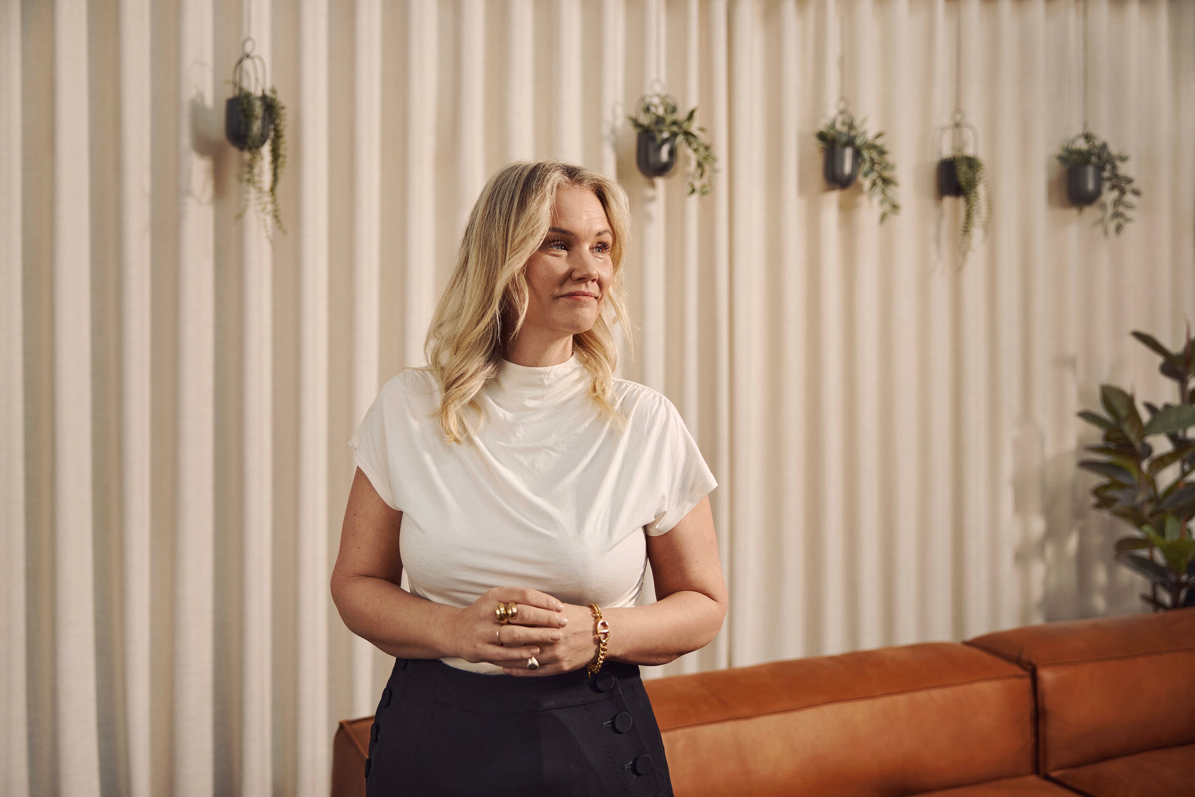 Woman in white t-shirt and dark skirt standing in front of striped wall with hanging plants