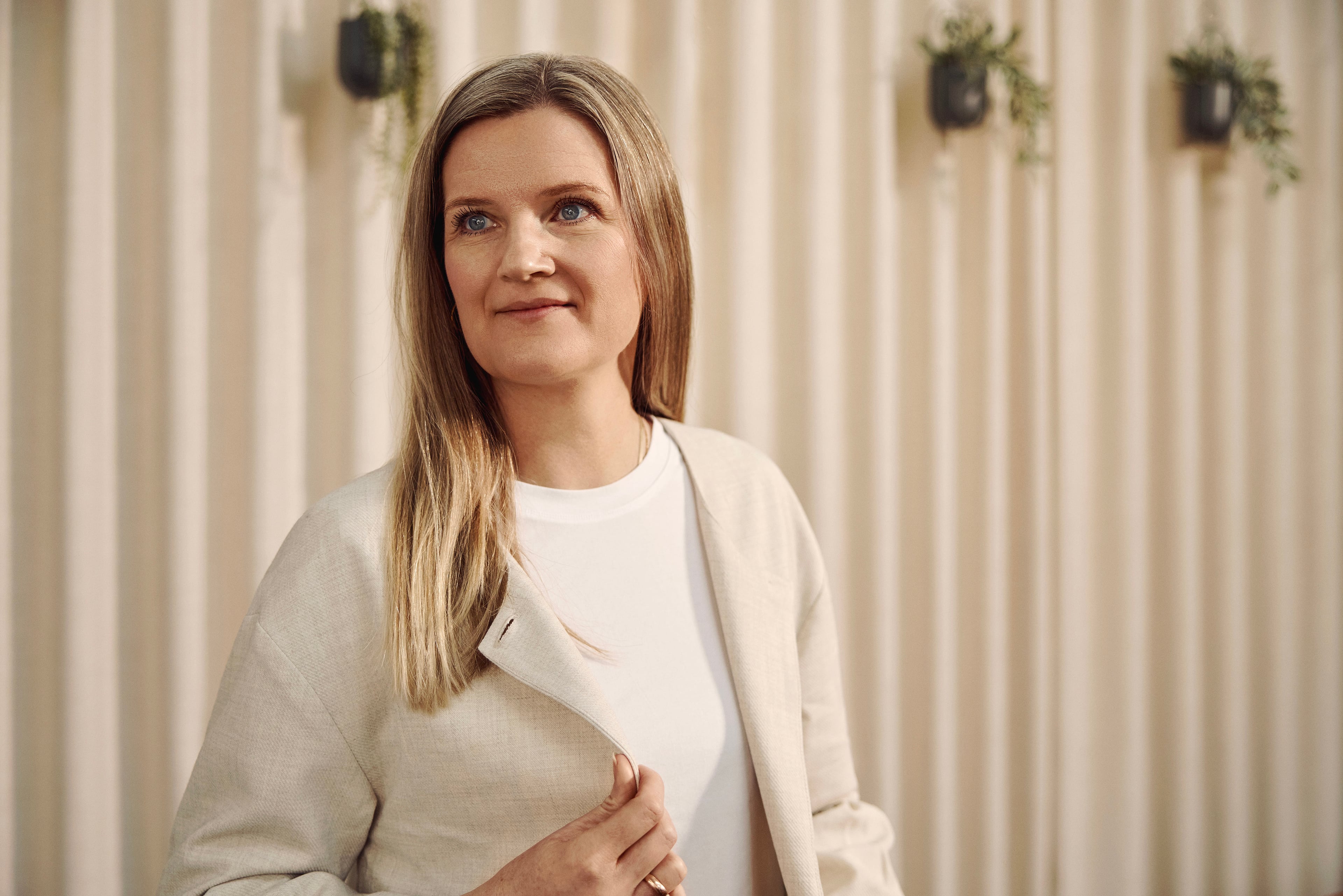 Woman in beige cardigan and white t-shirt standing in front of striped wall with hanging plants