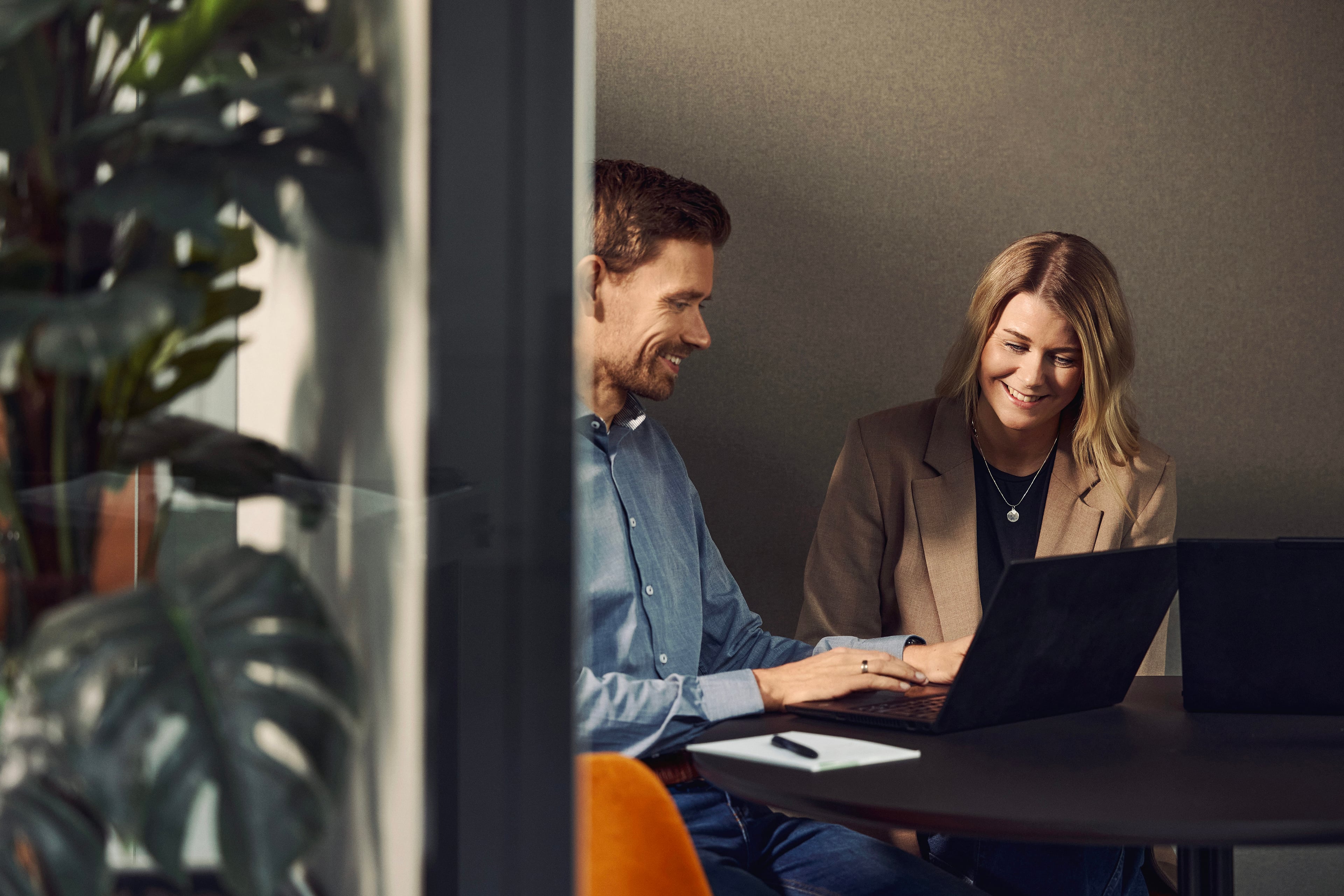 Two people working together at a laptop at a desk in an office with plants in the background.