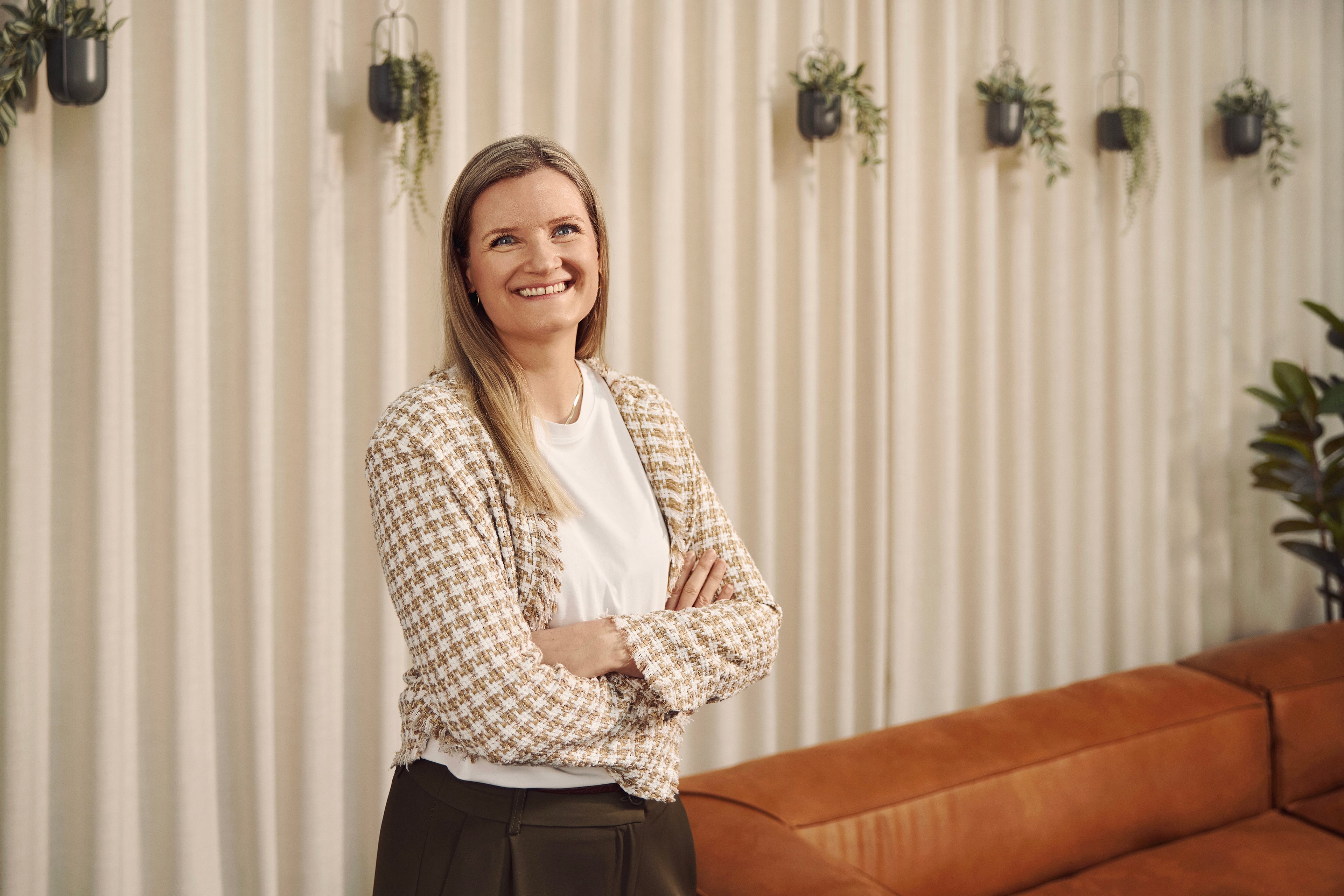 Woman in beige patterned blazer standing in front of striped wall with hanging plants and leather sofa