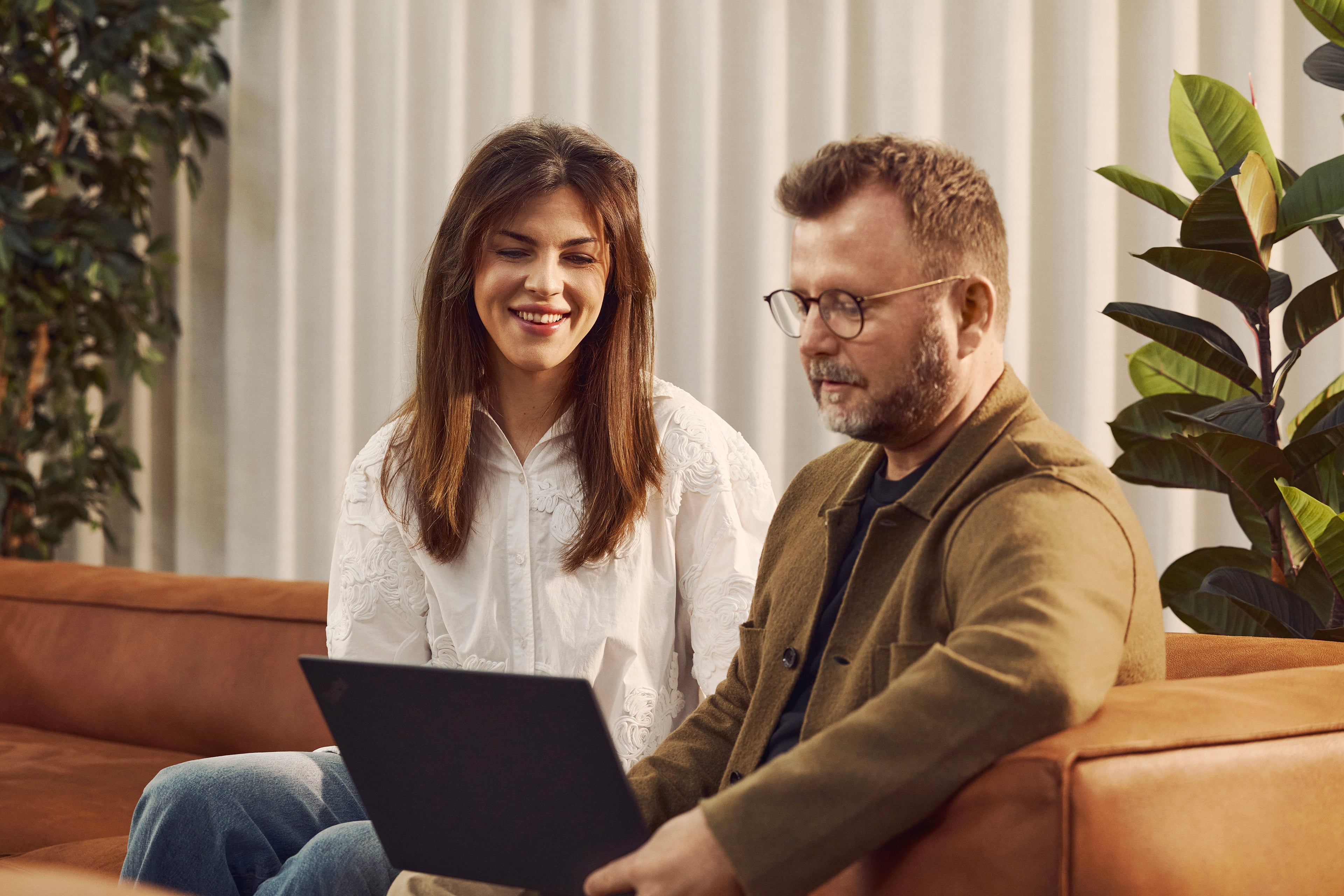 Two people sit on a brown sofa working together on a laptop. Woman in white shirt and man in brown jacket.