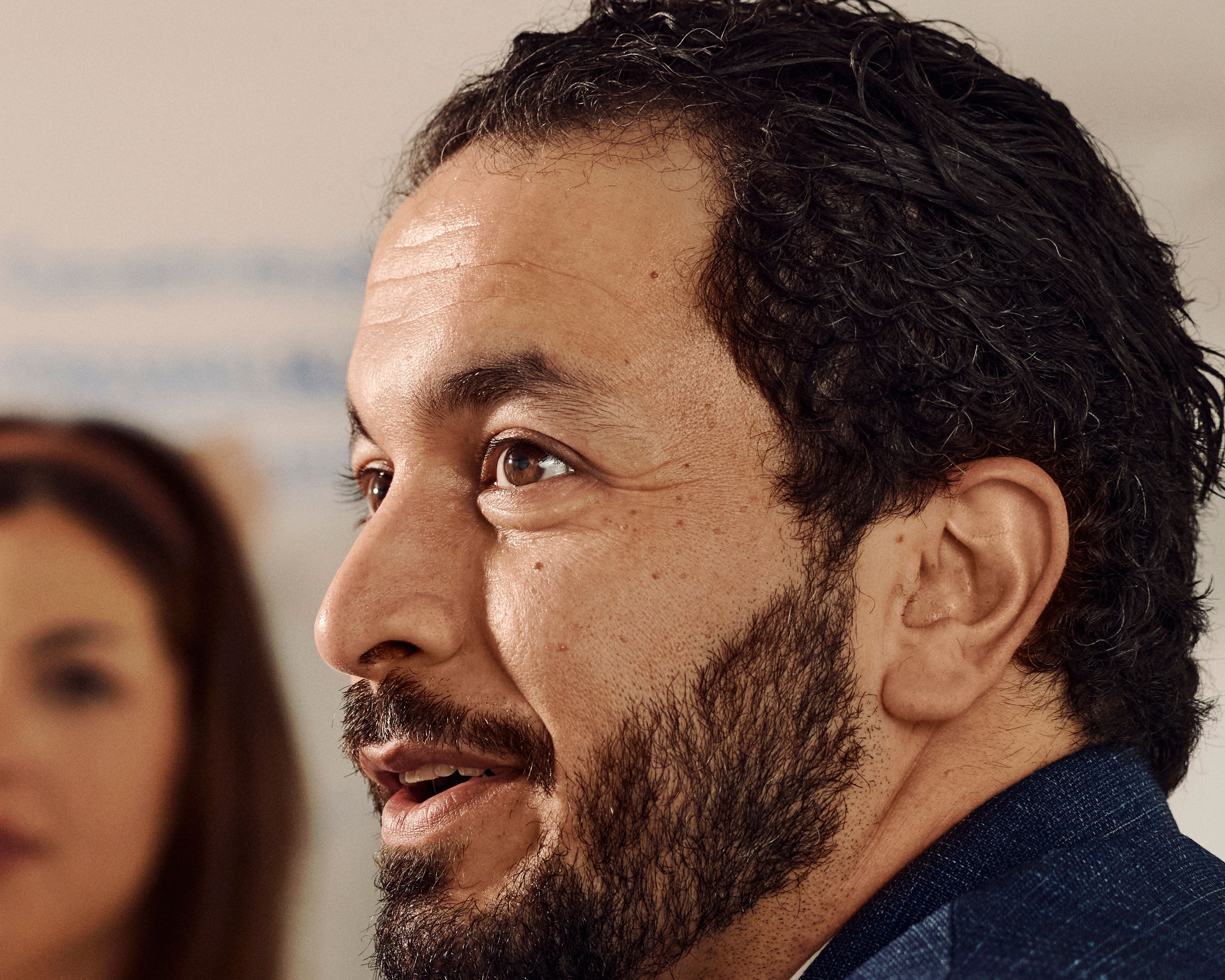 Bearded man in profile looking to the side with expressive face. Another person blurred in background.